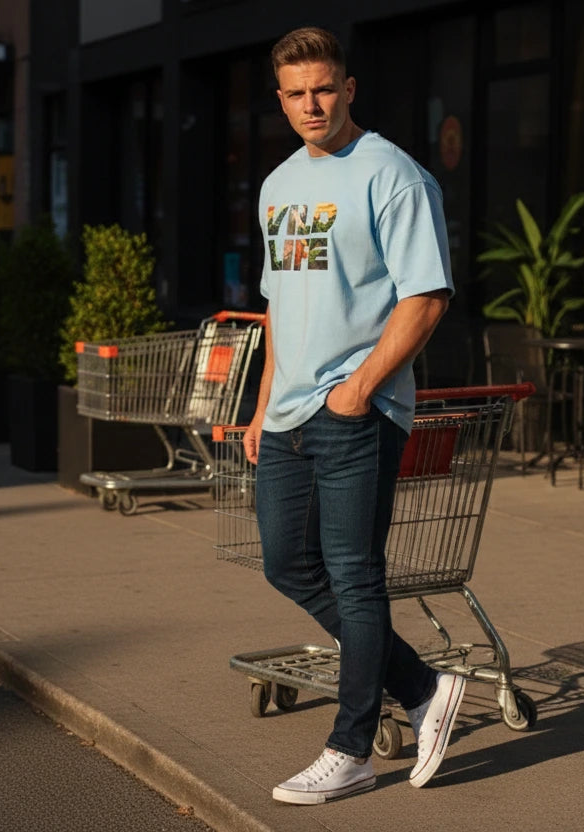 Man standing on a sidewalk with shopping carts, wearing a light blue t-shirt and dark jeans.