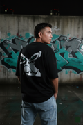 Person wearing a black t-shirt with a white graphic design in front of a graffiti-covered wall.