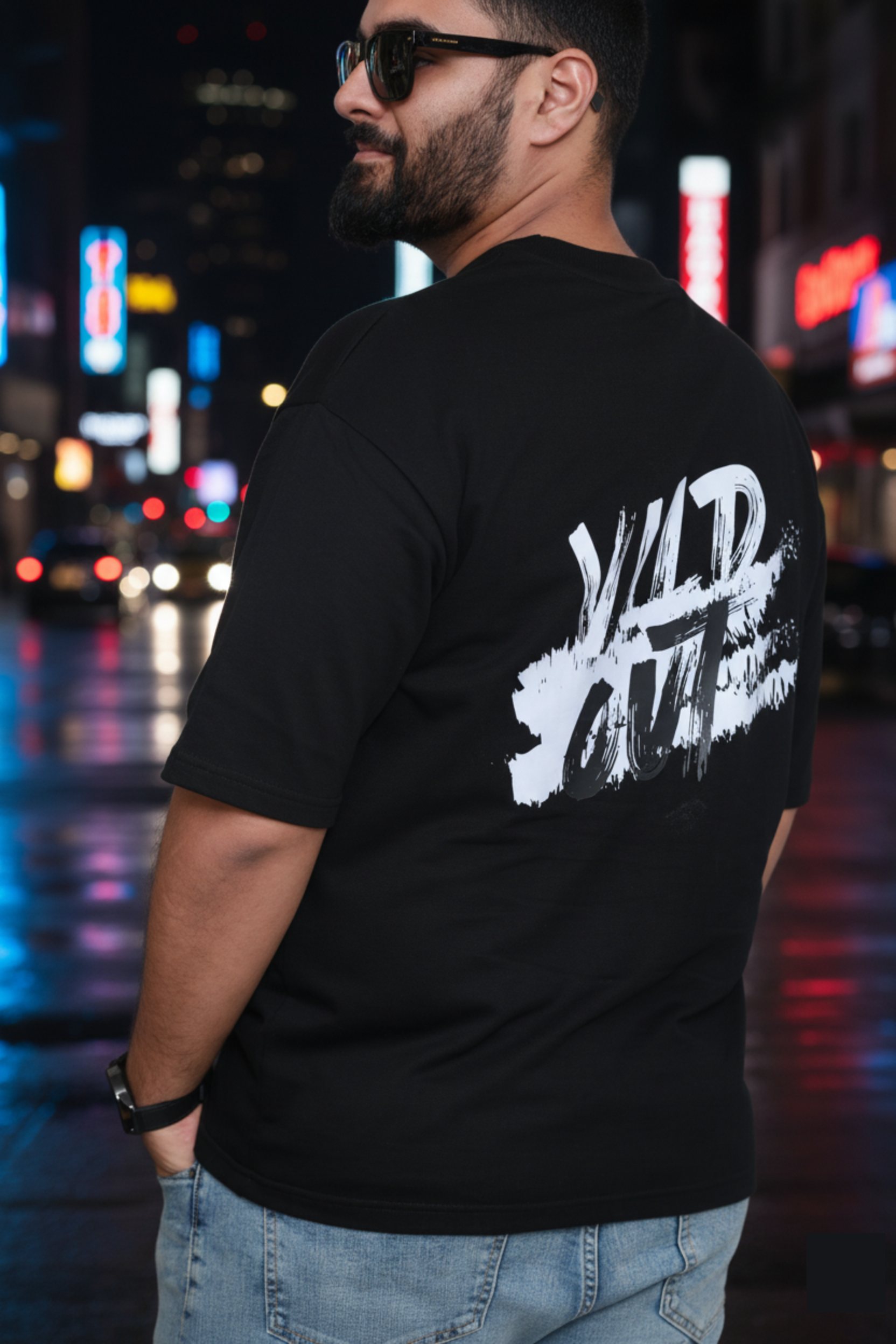 Man wearing a black t-shirt with white graphic design on the back, standing in an urban night setting with neon lights.