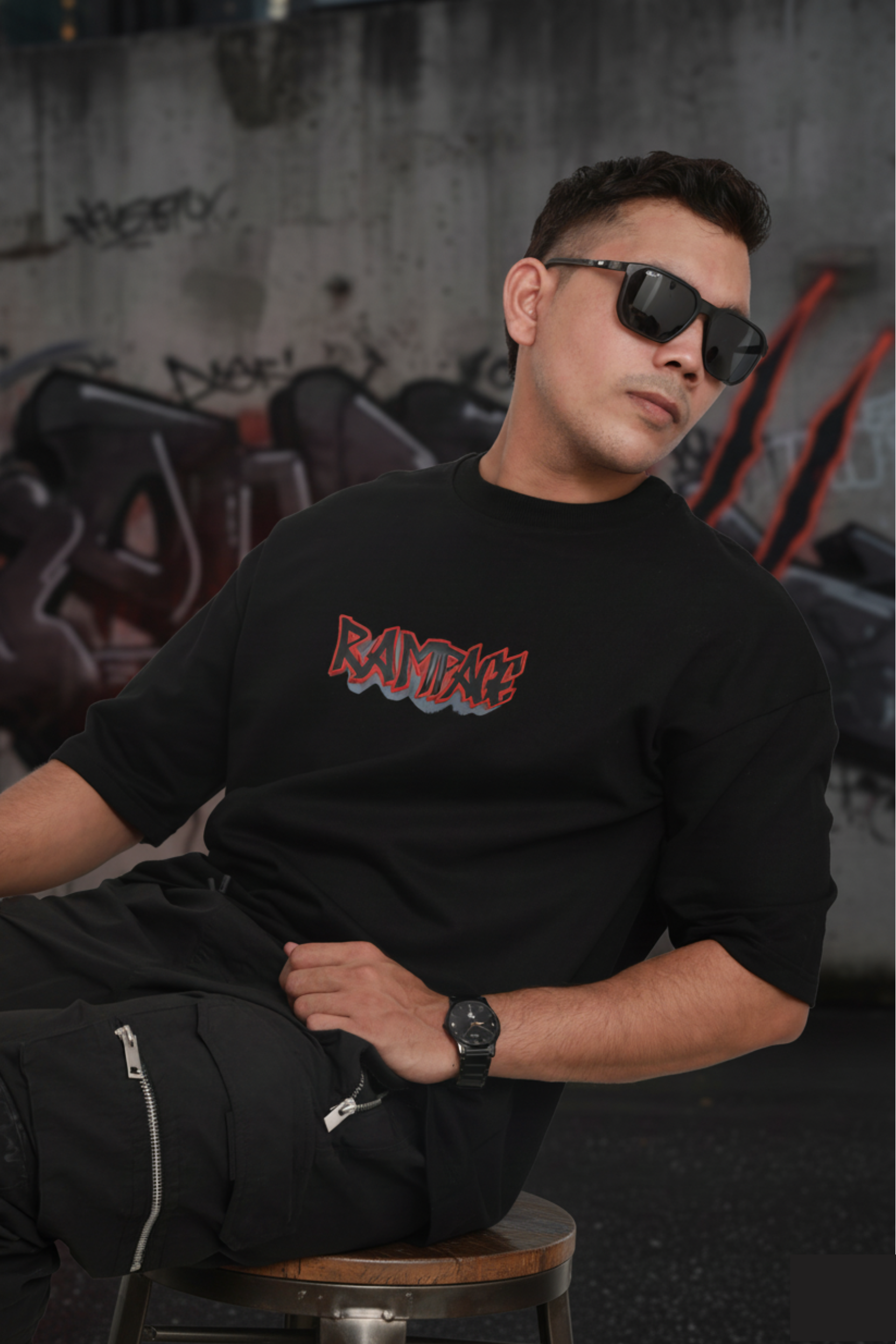Man wearing a black t-shirt with colorful text design, sitting against a graffiti-covered wall.