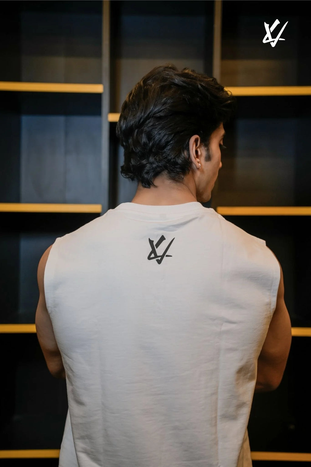Man wearing a sleeveless shirt with a logo on the back, standing in front of a dark wall with horizontal lines.